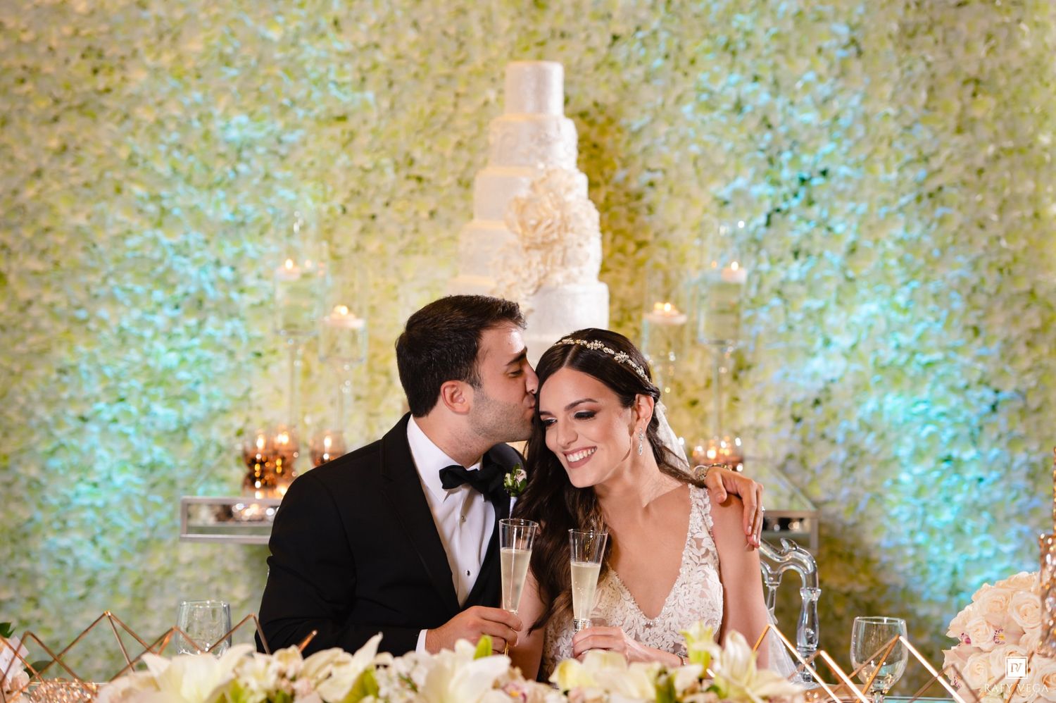 Bride and groom share a romantic moment at wedding reception with white floral wall backdrop and elegant tiered cake.