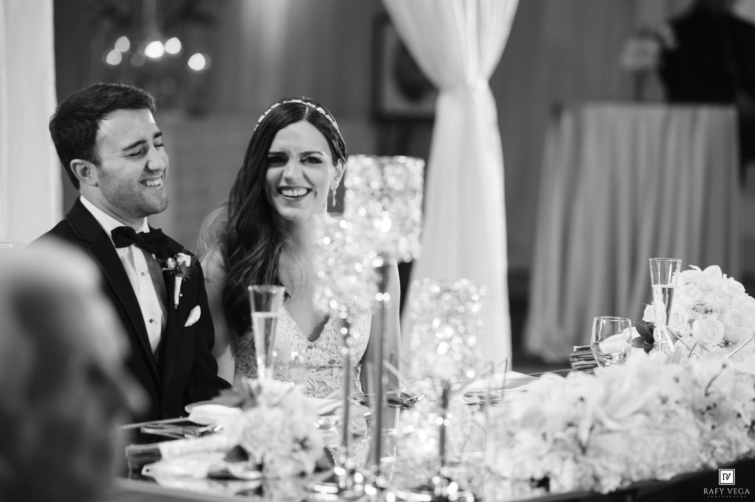 Elegant black and white wedding reception photo of smiling bride in sequin gown and groom in tuxedo seated at floral head table.