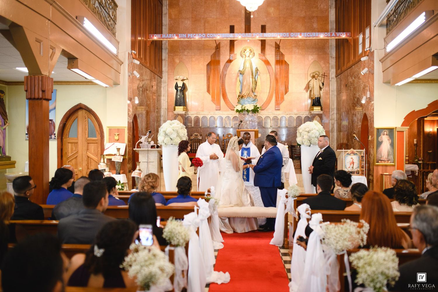 A couple exchanges wedding vows at a decorated church altar with white floral arrangements and guests seated in pews.