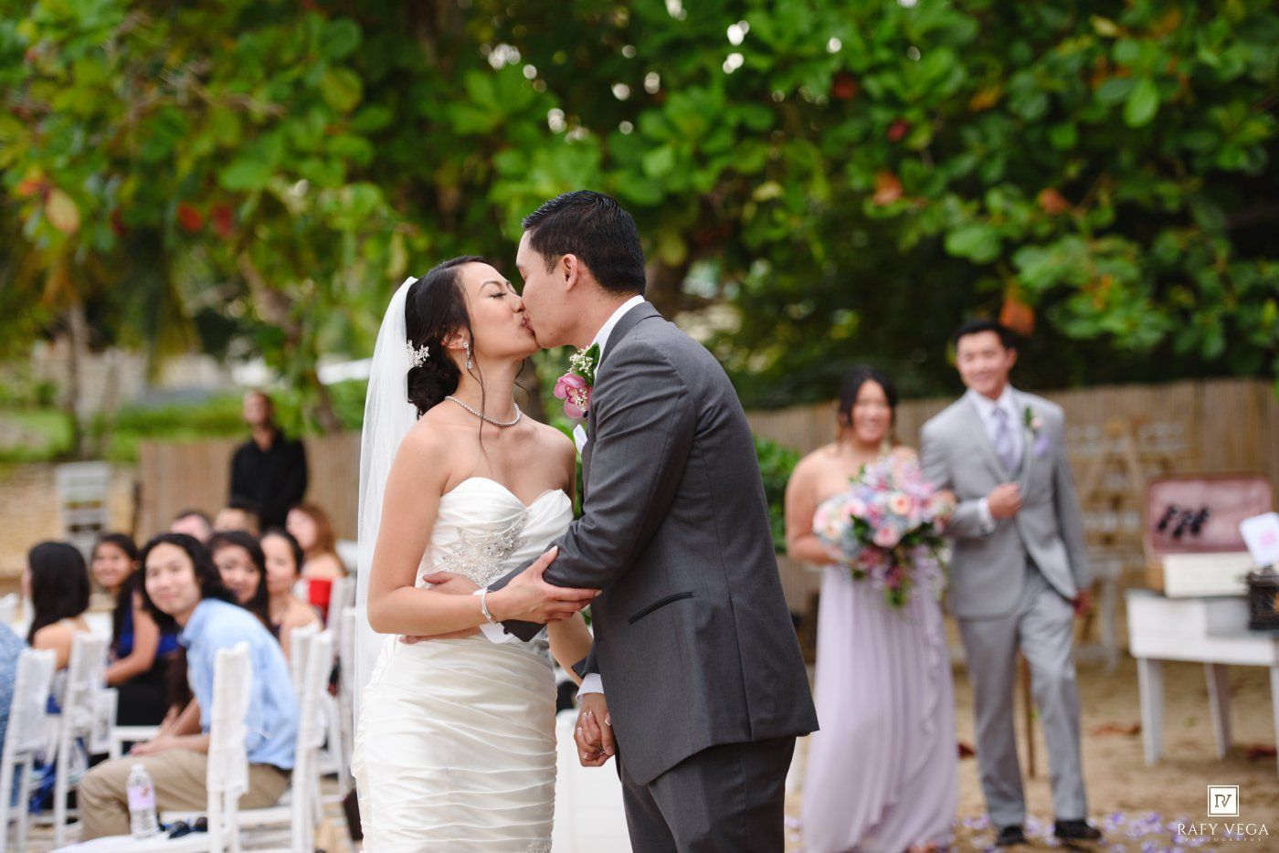 Bride and groom share first kiss at outdoor wedding ceremony with guests seated in background.