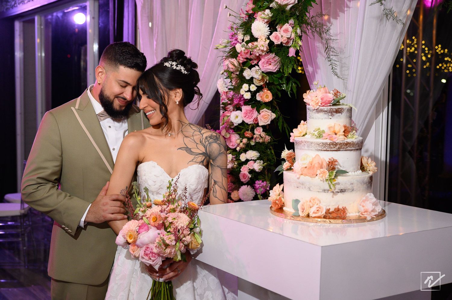 Bride in a white gown holding a pink bouquet stands beside a groom in an olive suit next to a tiered wedding cake and floral arrangement.