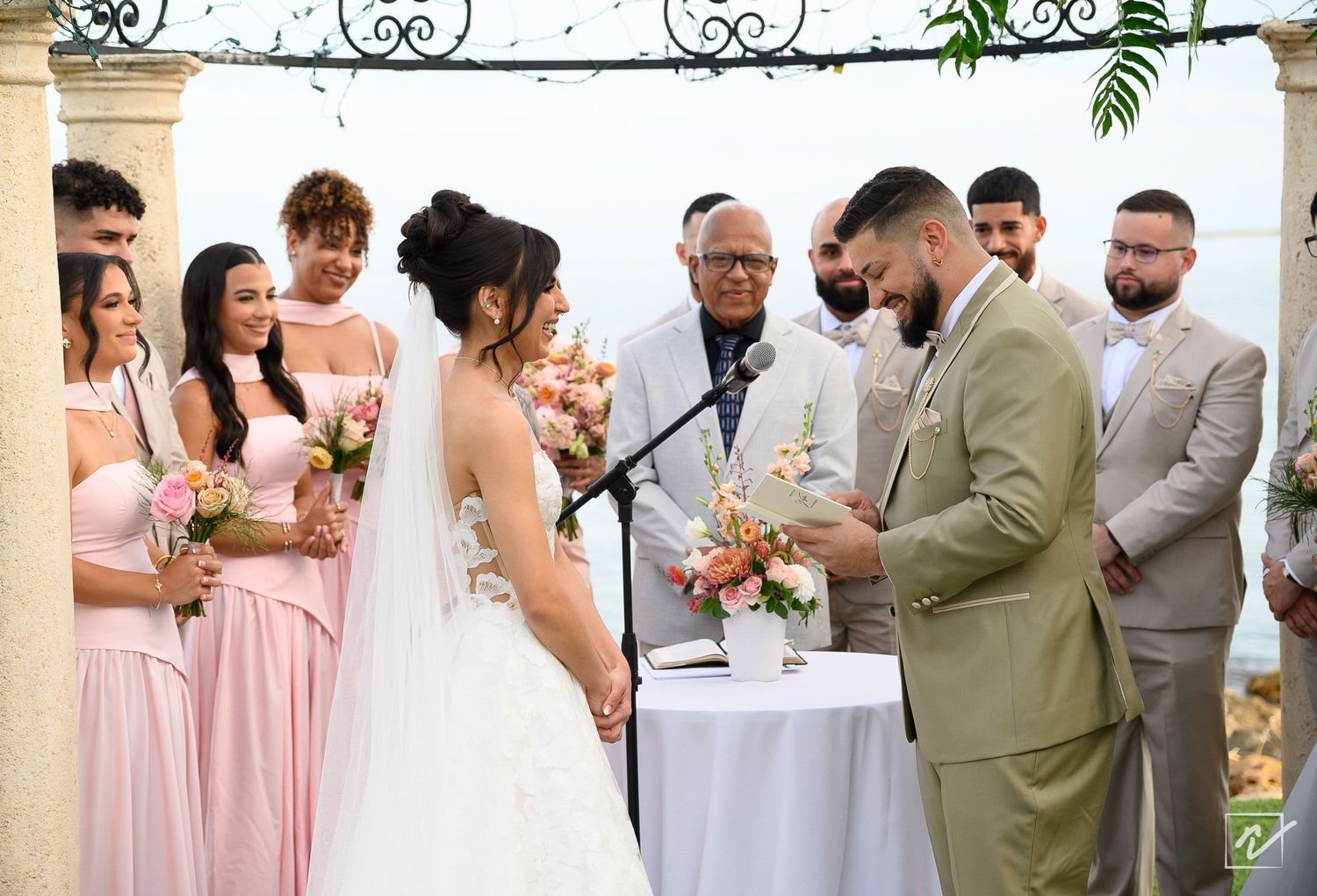 Bride in a white gown and groom in an olive suit exchange vows at an outdoor wedding as bridesmaids in blush dresses and groomsmen watch.