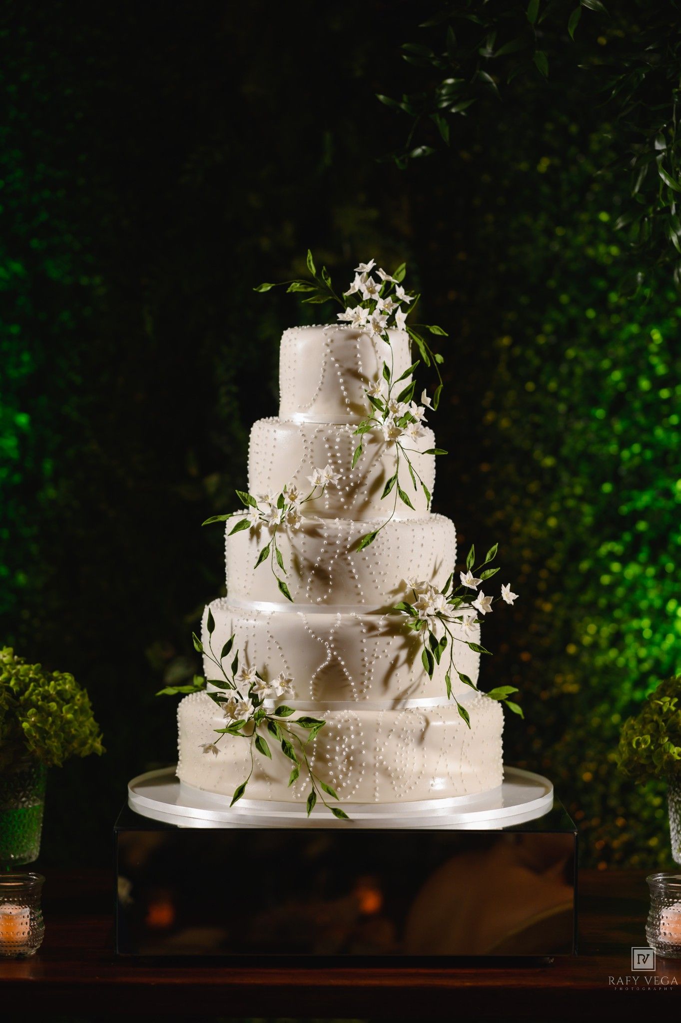 Elegant five-tier white wedding cake adorned with pearl details and fresh greenery branches against a lush green backdrop.