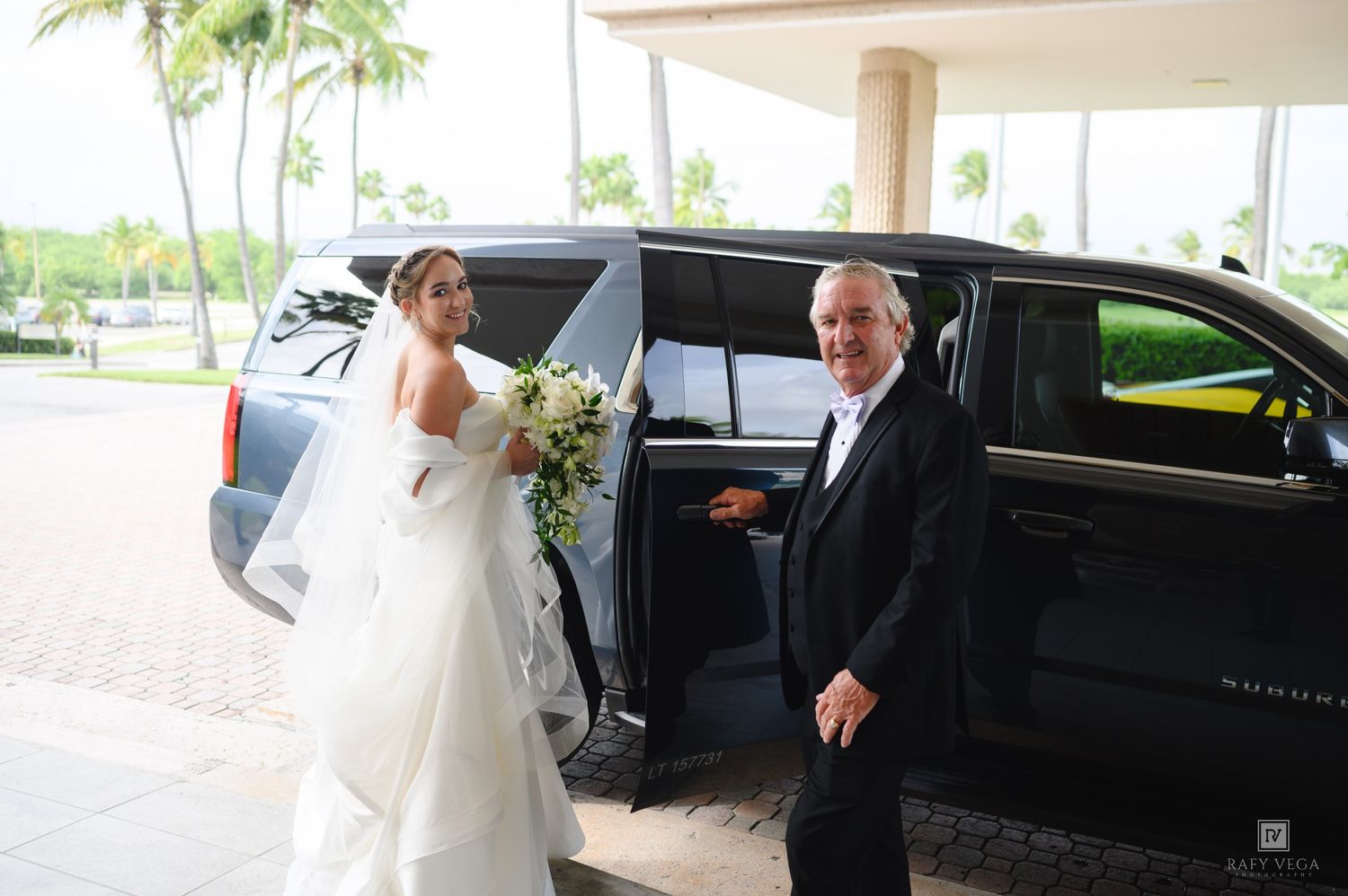Bride in white gown holding bouquet exits black SUV limo assisted by man in tuxedo at tropical wedding venue.