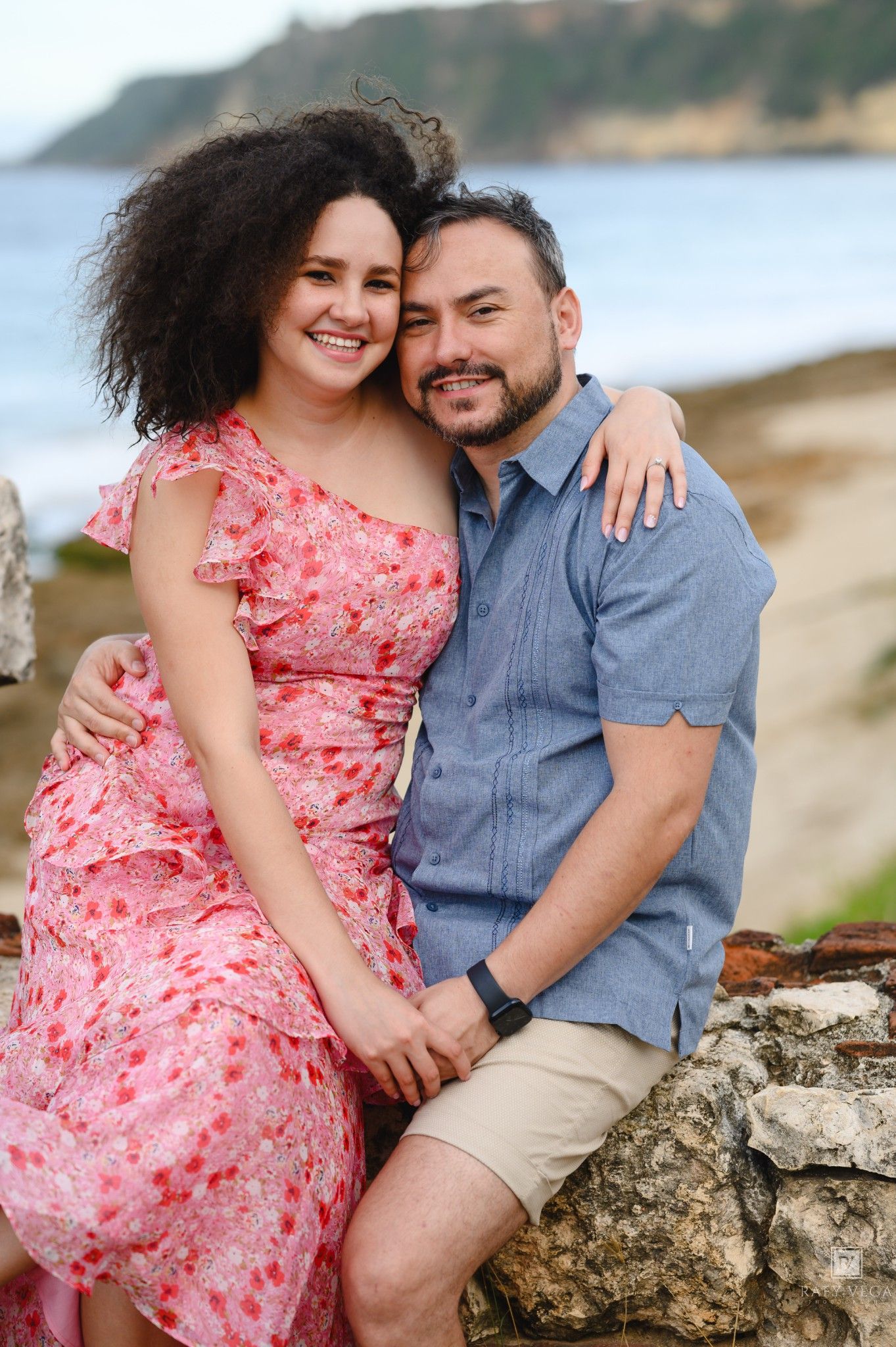 A smiling couple embraces on a rocky seaside wall, woman in pink floral dress, man in blue shirt, ocean backdrop.