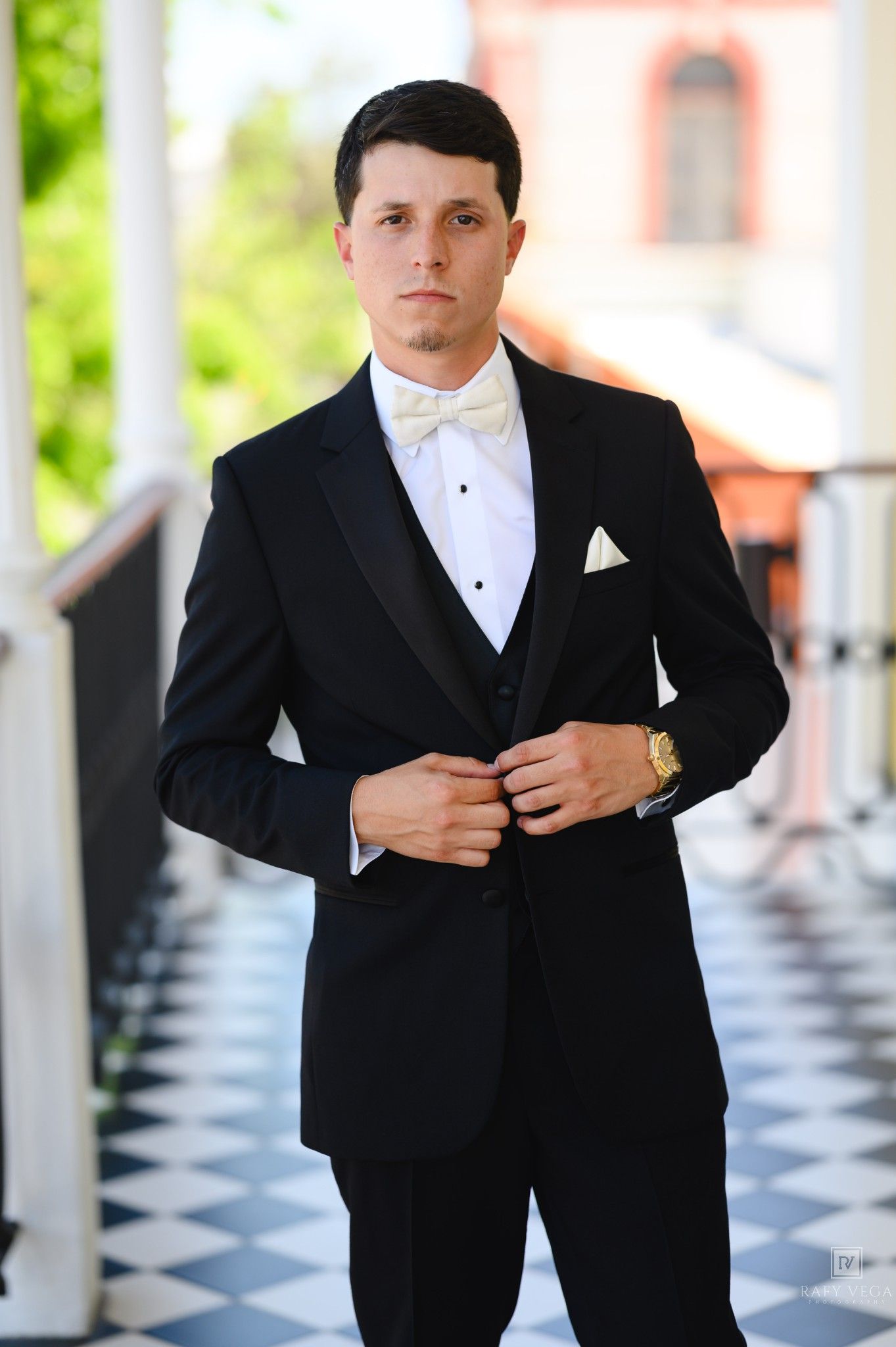 Elegant groom in a black tuxedo with ivory bow tie and pocket square poses confidently at a wedding venue.