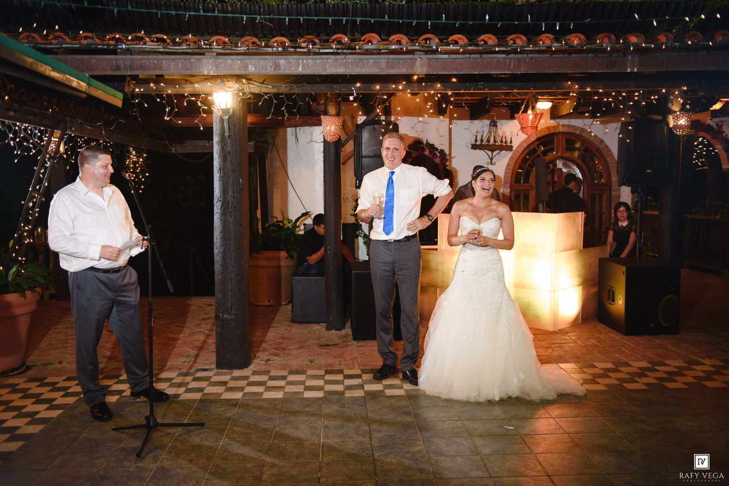 Bride and groom share a joyful moment during wedding reception toast in a warmly lit venue with string lights.