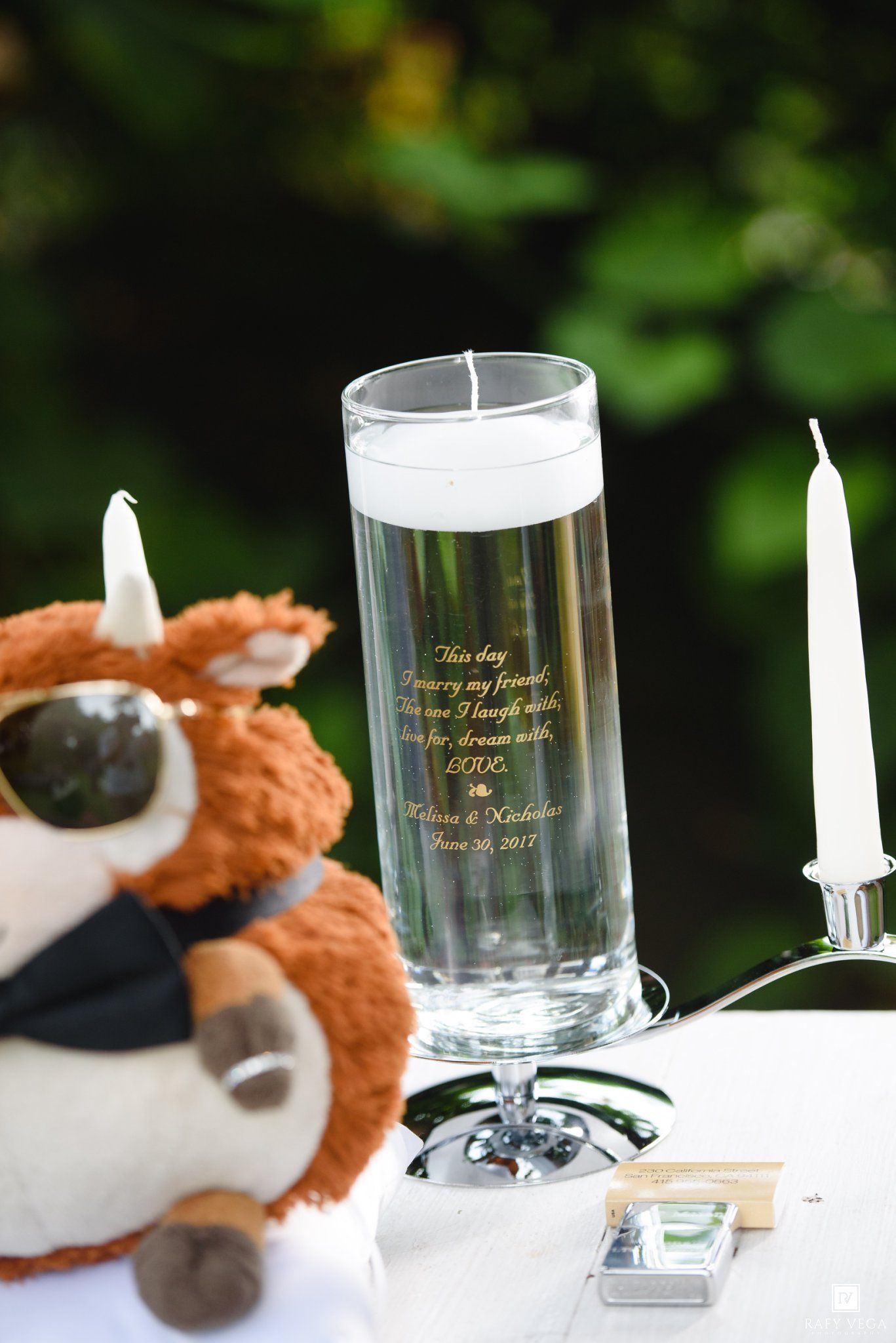 Personalized wedding unity candle vase engraved with vows, flanked by white taper candles and a stuffed animal toy.