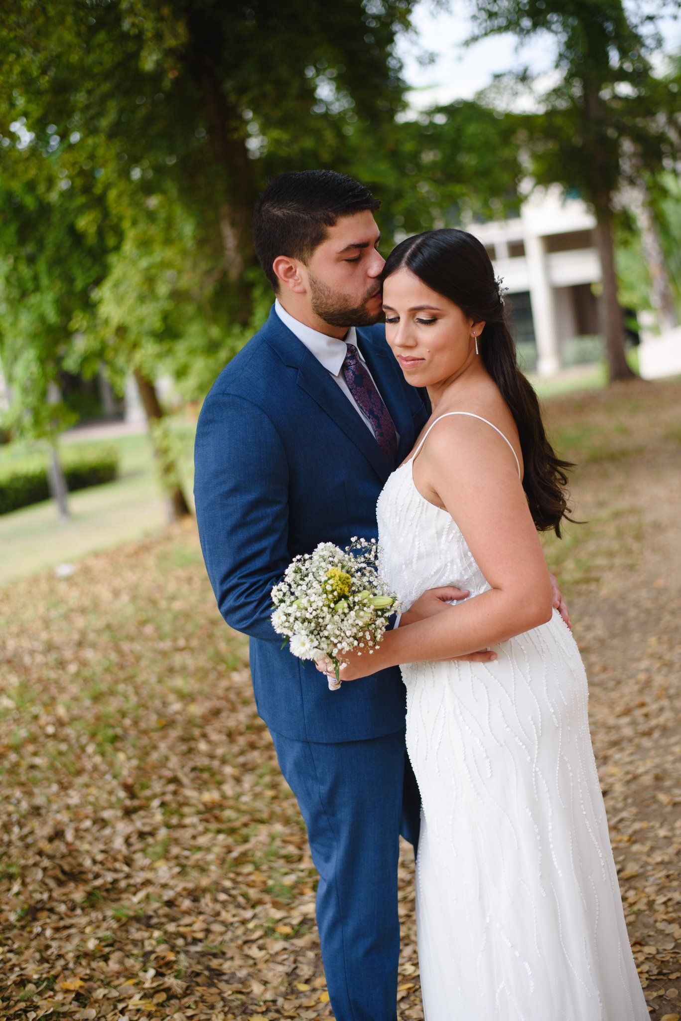 Groom in blue suit kissing bride in white dress holding wildflower bouquet in a tree-lined park setting.