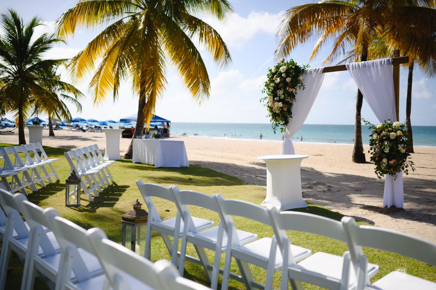 Elegant beachside wedding setup with white chairs, floral arch, and palm trees on a tropical sandy shore.