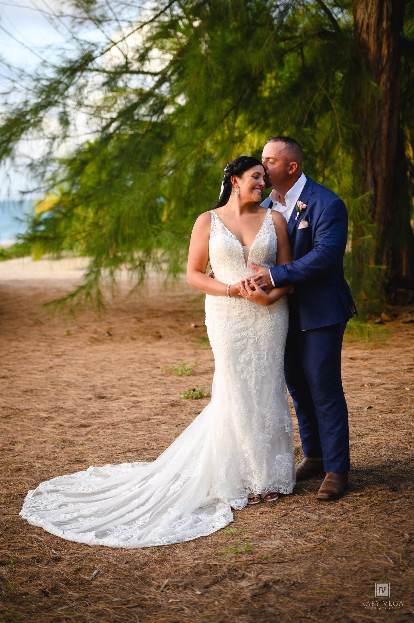 Bride and groom share a romantic embrace under pine trees on a sandy beach, bride wearing a lace mermaid gown.
