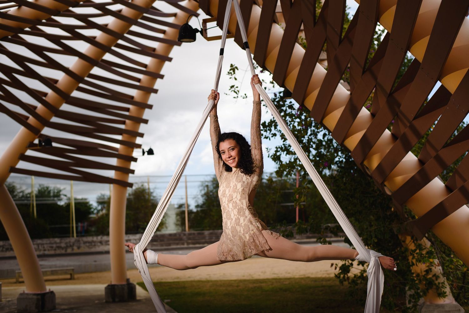 A flexible dancer performs a split pose on aerial silks under a wooden lattice structure in an outdoor park setting.