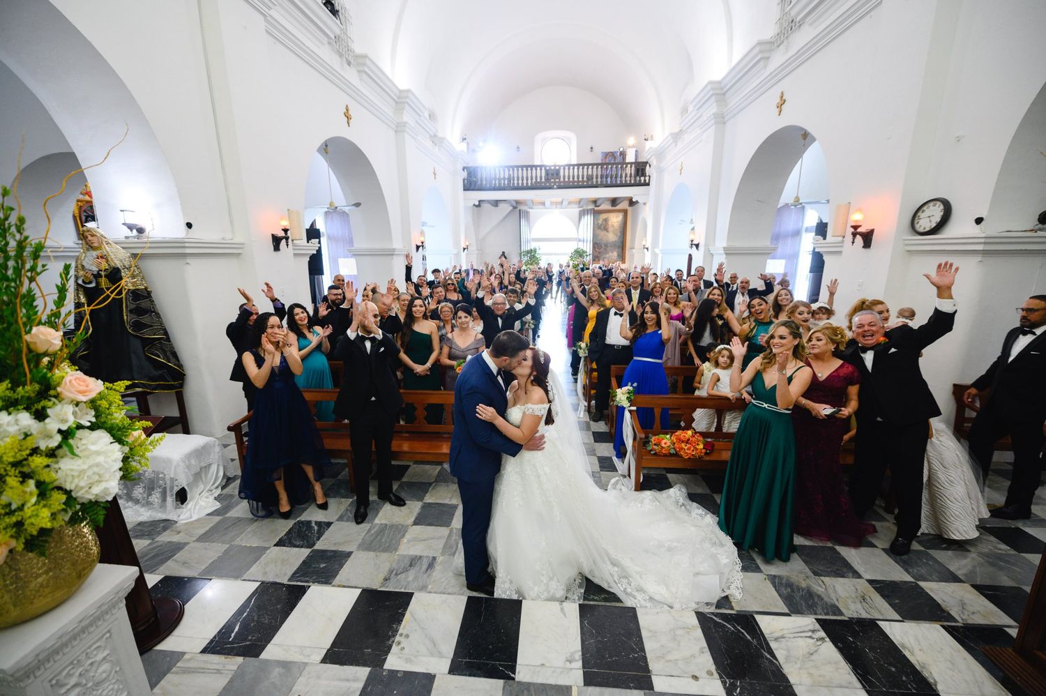 Joyful wedding ceremony in elegant white church with bride and groom kissing surrounded by cheering guests.