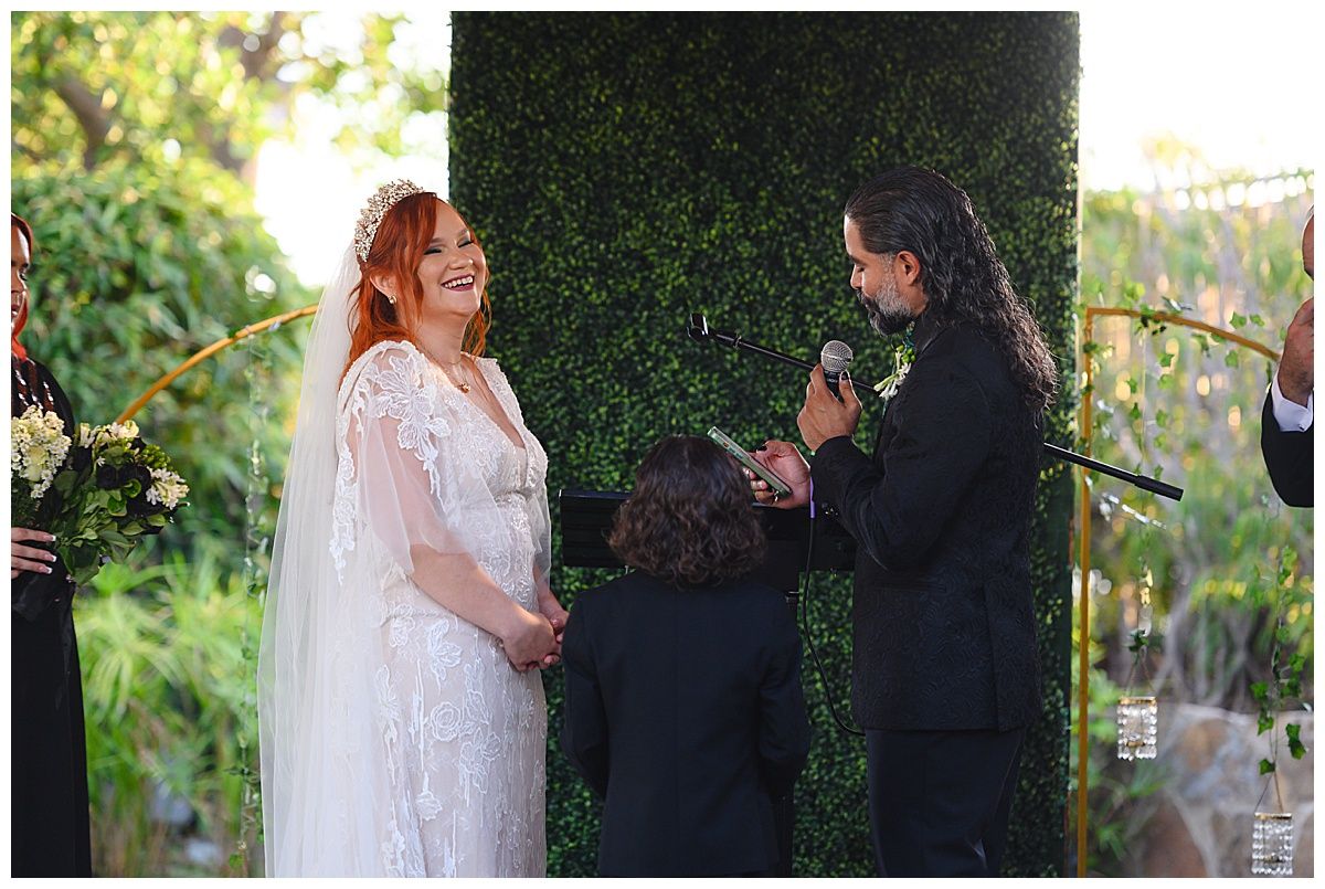 Red-haired bride laughs joyfully as groom speaks during outdoor garden wedding ceremony.