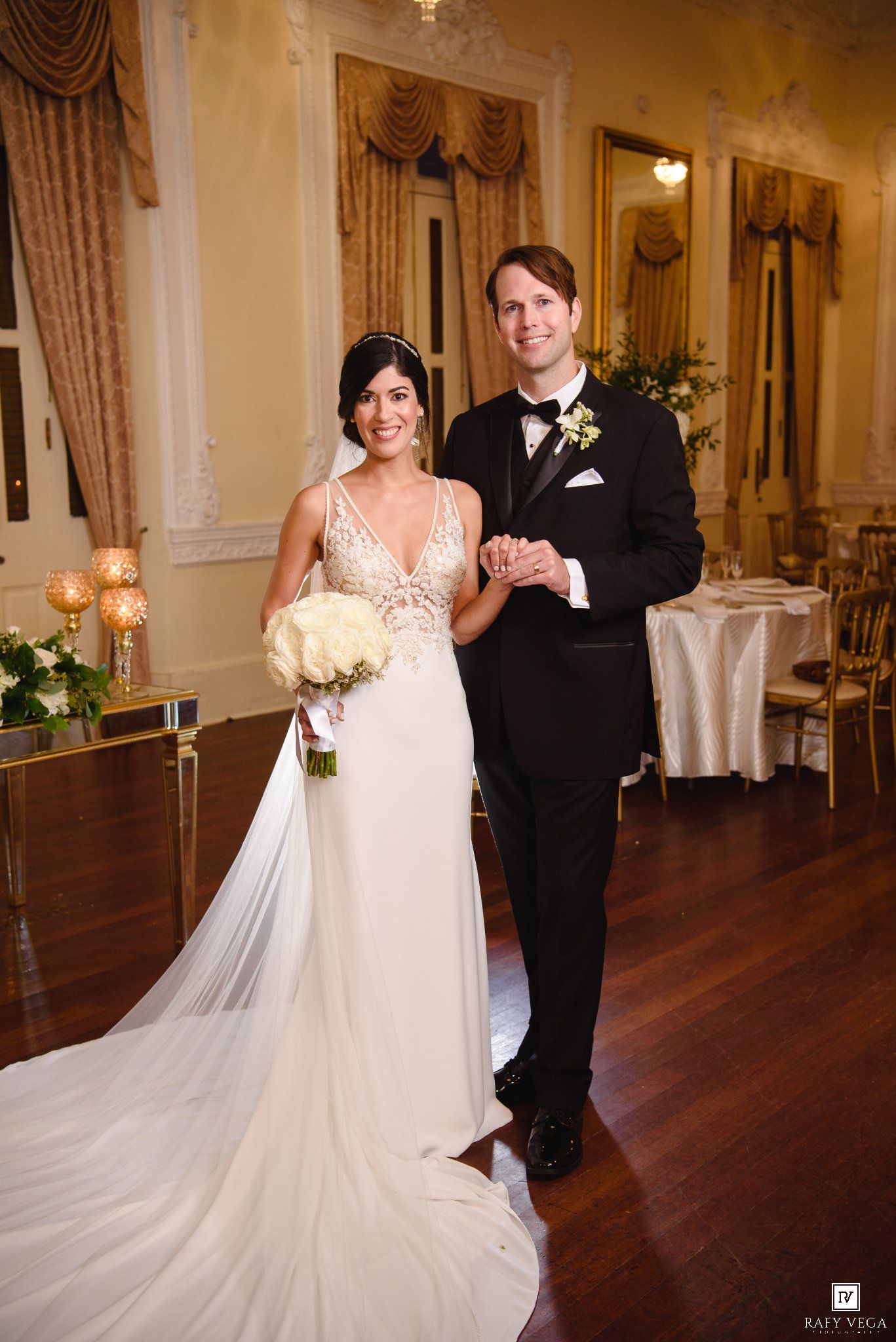 Bride in white gown holding bouquet stands with groom in tuxedo in elegant ballroom wedding venue.