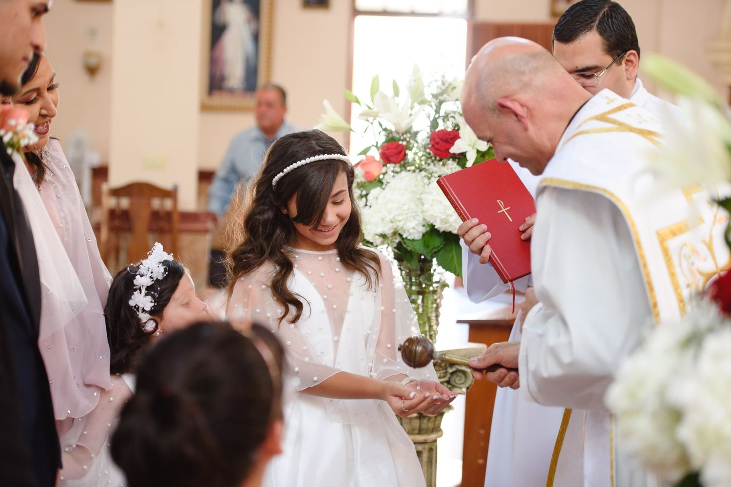 Girl in white dress receives First Communion from priest in church ceremony with floral arrangements.