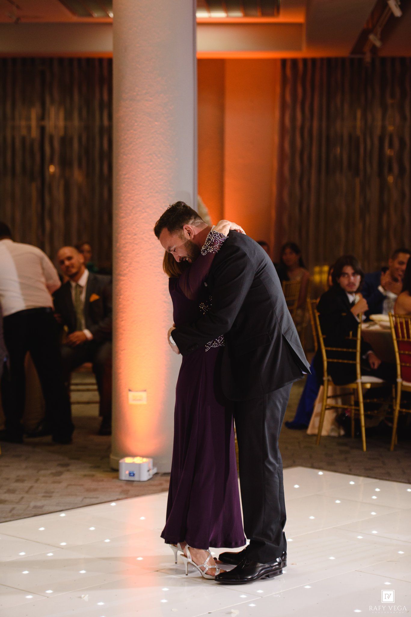 Couple sharing an intimate dance on white dance floor at elegant wedding reception with warm amber lighting.