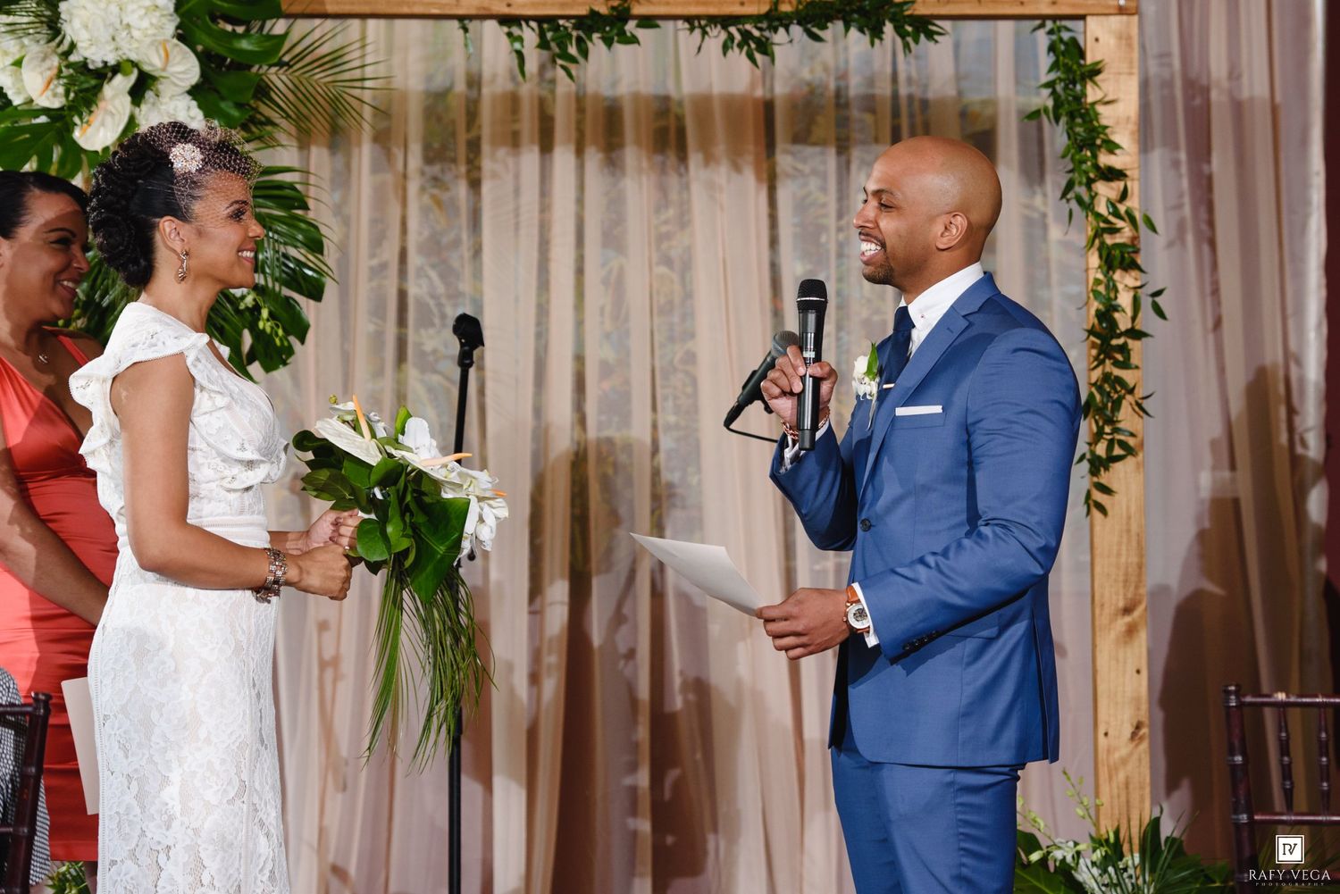 Groom in blue suit reads wedding vows to bride in white dress at elegant floral altar ceremony.