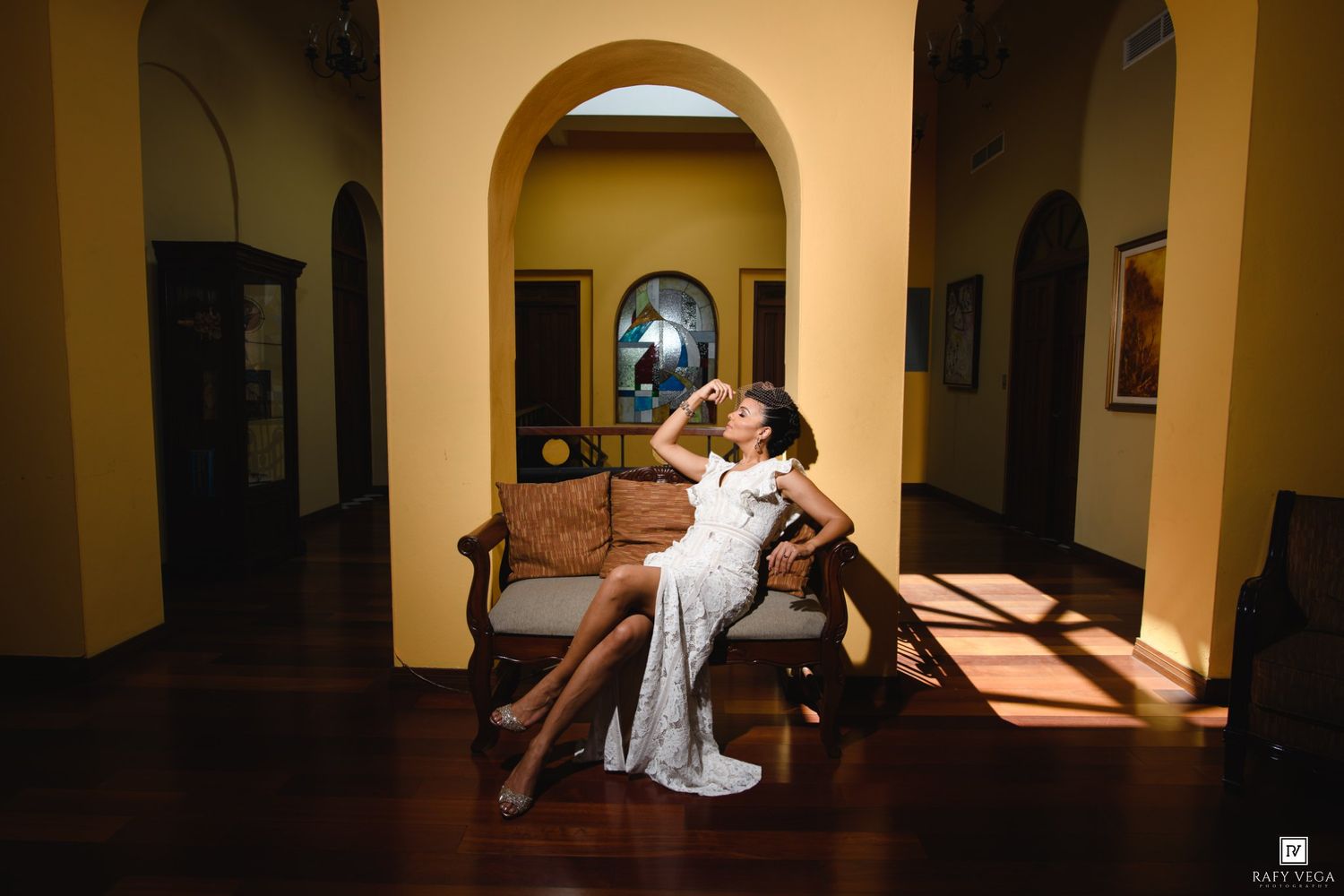 Couple in elegant wedding attire sharing a romantic moment on a wooden bench in a sunlit colonial hallway.