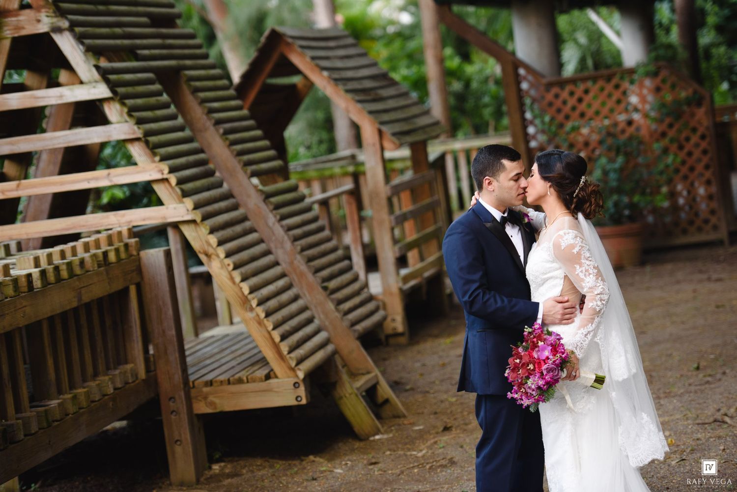 A bride and groom share a romantic kiss outdoors near wooden structures, bride holding a pink bouquet in a lace wedding gown.