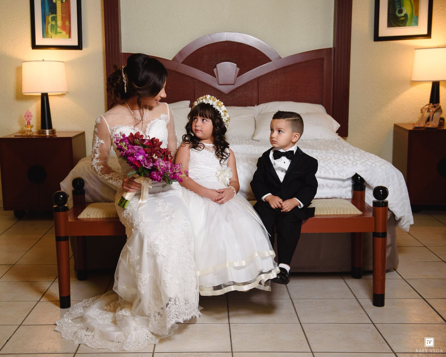 Bride in lace wedding gown sits on bed with flower girl and ring bearer in elegant hotel room.