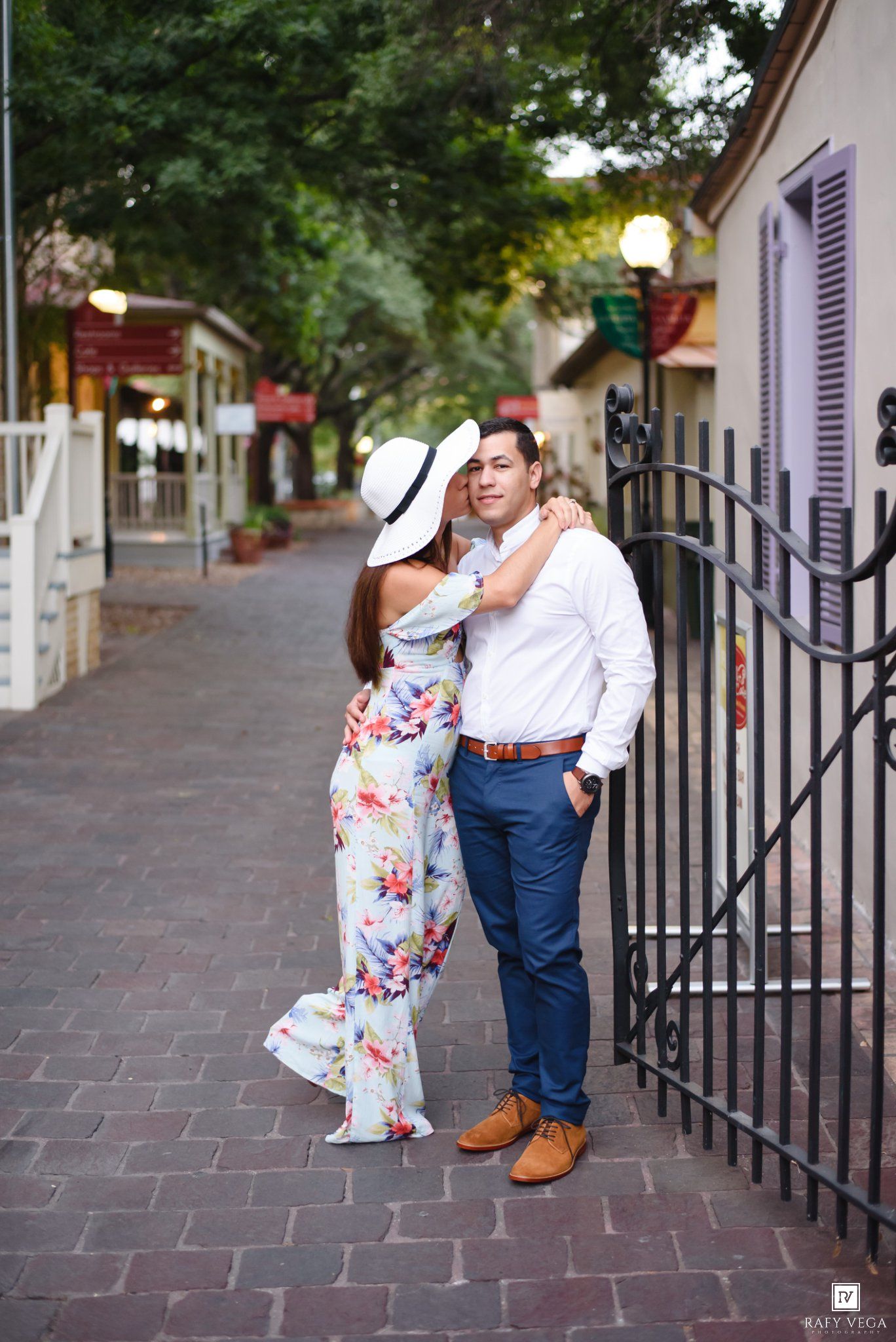 Romantic couple posing by black iron gate on quaint brick street, woman in floral maxi dress.