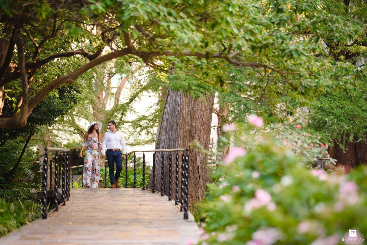 Couple walking on a bridge surrounded by lush green trees and pink flowers in a romantic garden setting.