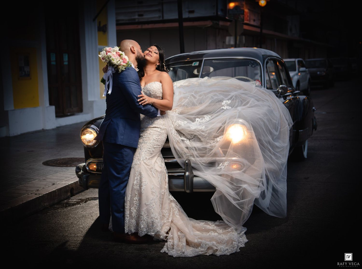 A bride and groom share a romantic moment beside a vintage black car, her veil flowing dramatically in the wind.