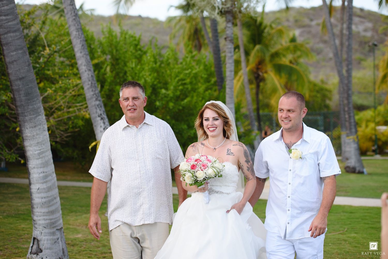Bride in white gown walks with two men in casual attire on tropical lawn with palm trees at outdoor wedding.