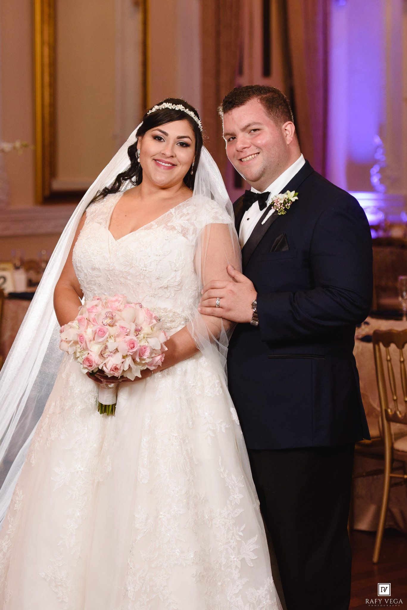 Bride and groom posing together at wedding reception, bride in lace gown holding pink bouquet, groom in navy tuxedo.