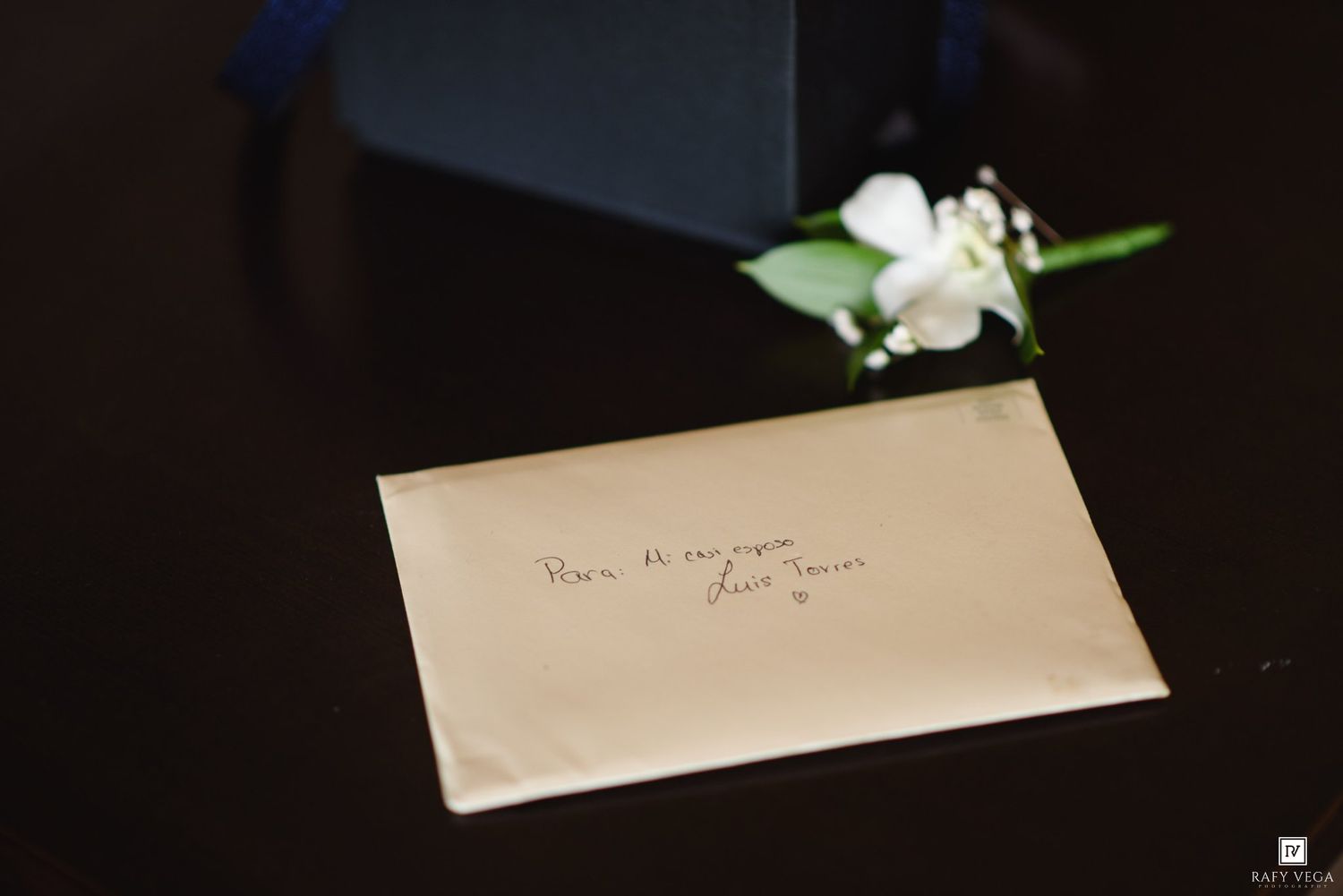 Wedding envelope with handwritten note beside white boutonniere flower on dark table.