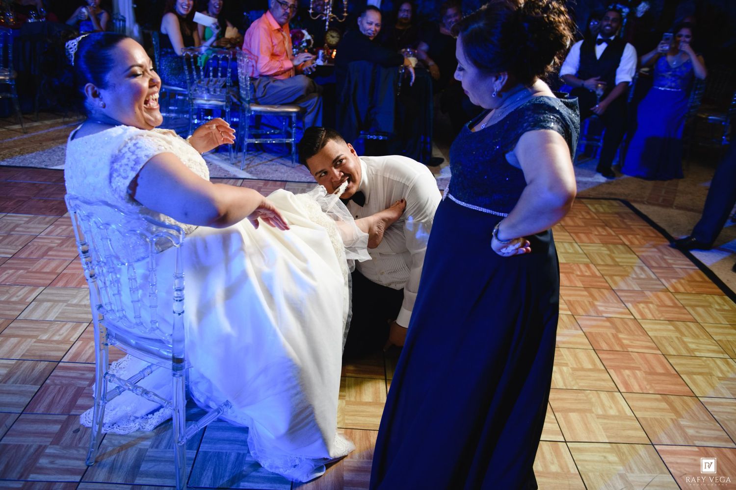 Groom kneeling before bride seated in chair on wedding dance floor, woman in blue gown watching nearby.