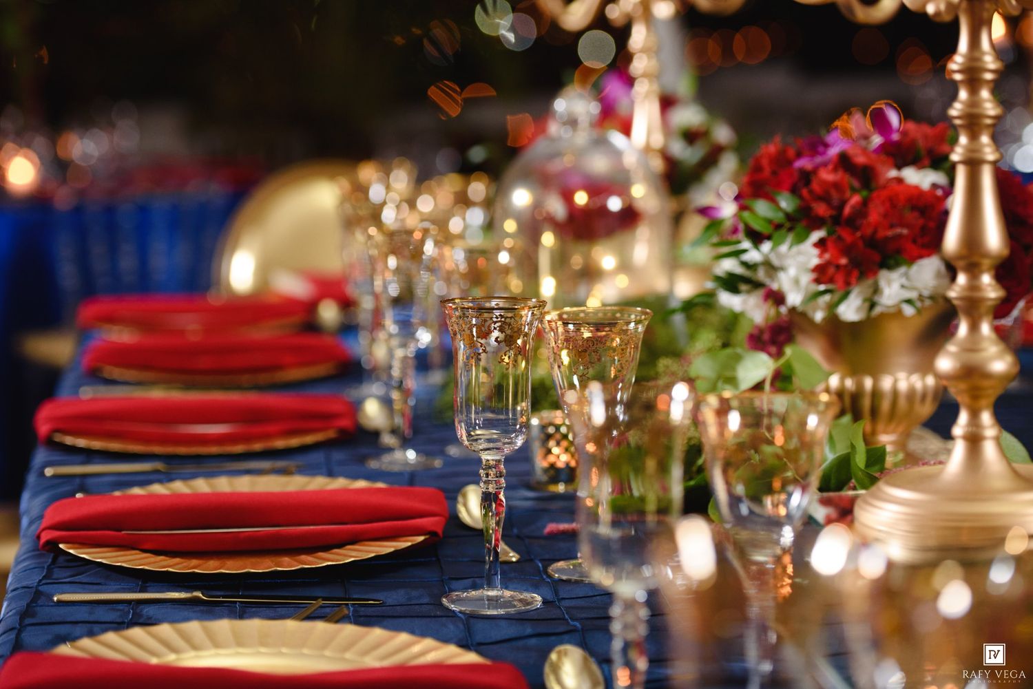 Elegant outdoor banquet table with blue velvet cloth, gold chargers, red plates, crystal glasses, and floral centerpieces.