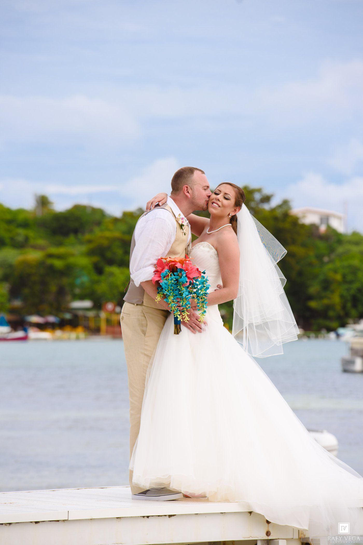 Bride and groom share a romantic kiss on a dock with tropical waterfront backdrop on their wedding day.