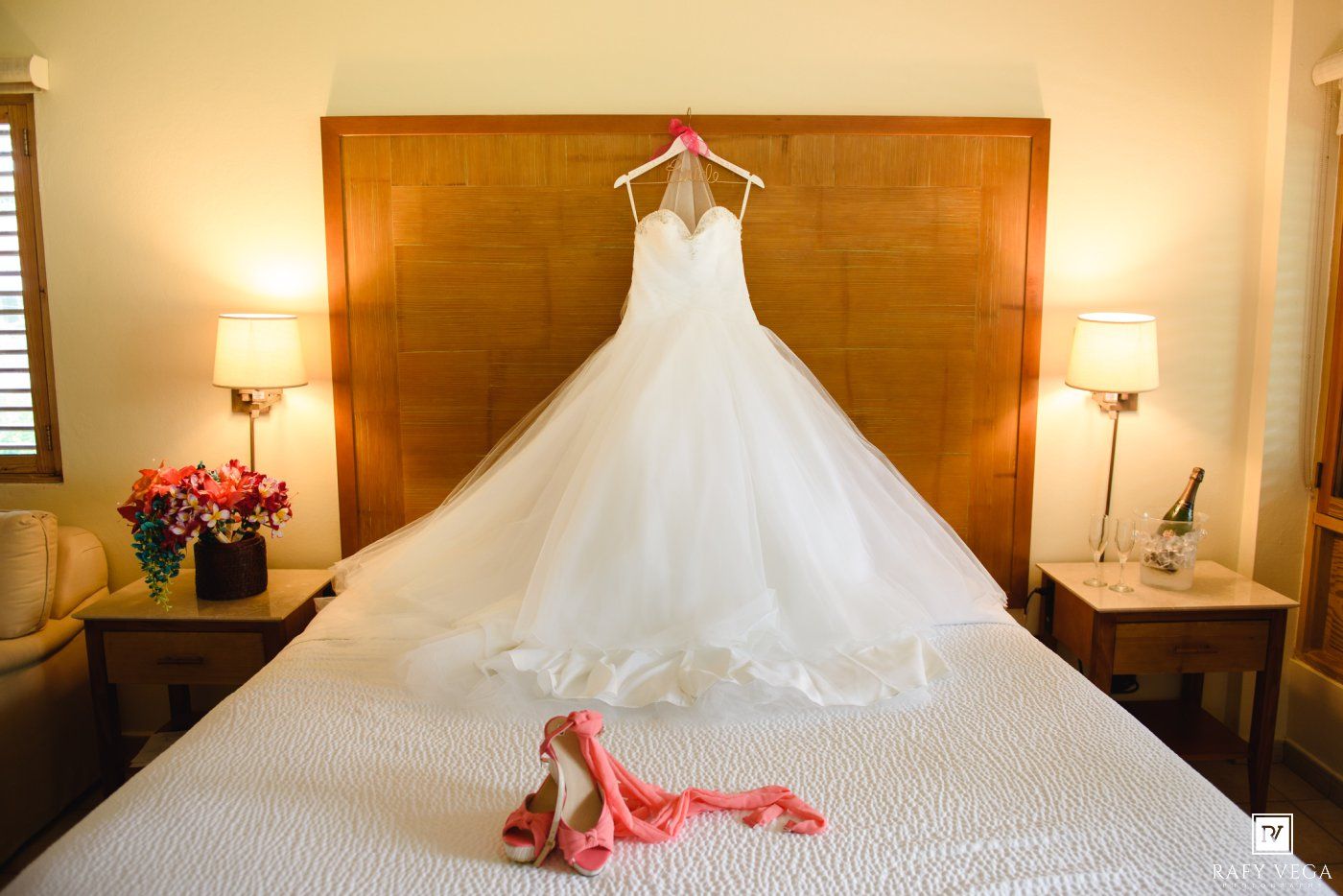 Elegant white ball gown wedding dress hanging above a bed with pink heels and sash in a warmly lit hotel room.