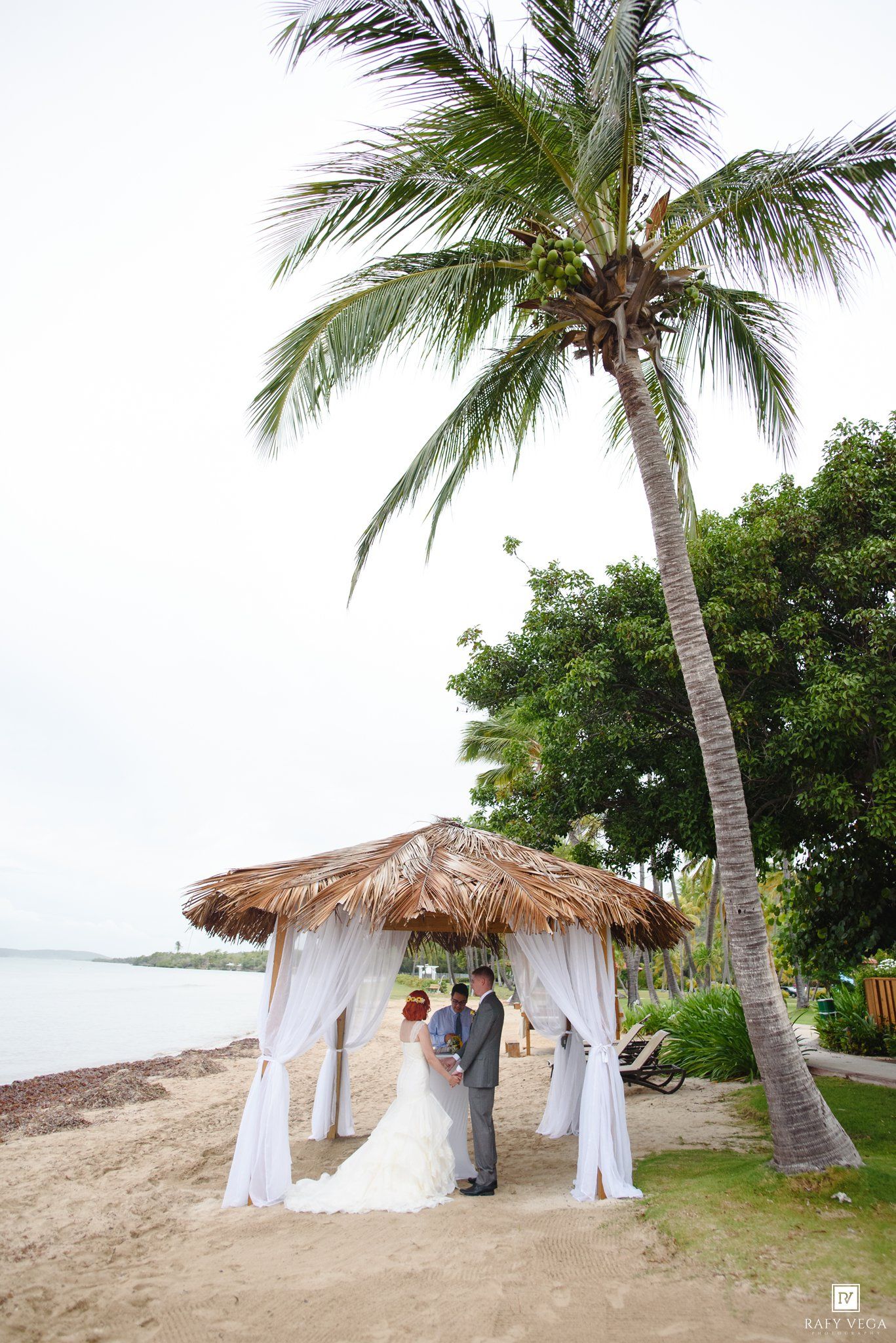 Couple exchanges vows under a thatched palapa on a tropical beach with palm trees during a destination wedding ceremony.