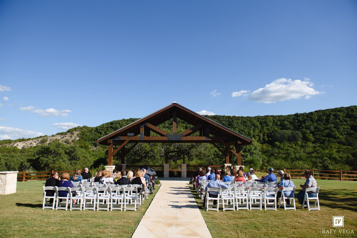 Outdoor wedding ceremony under a rustic pavilion with white chairs, lush green hills, and blue skies in the background.