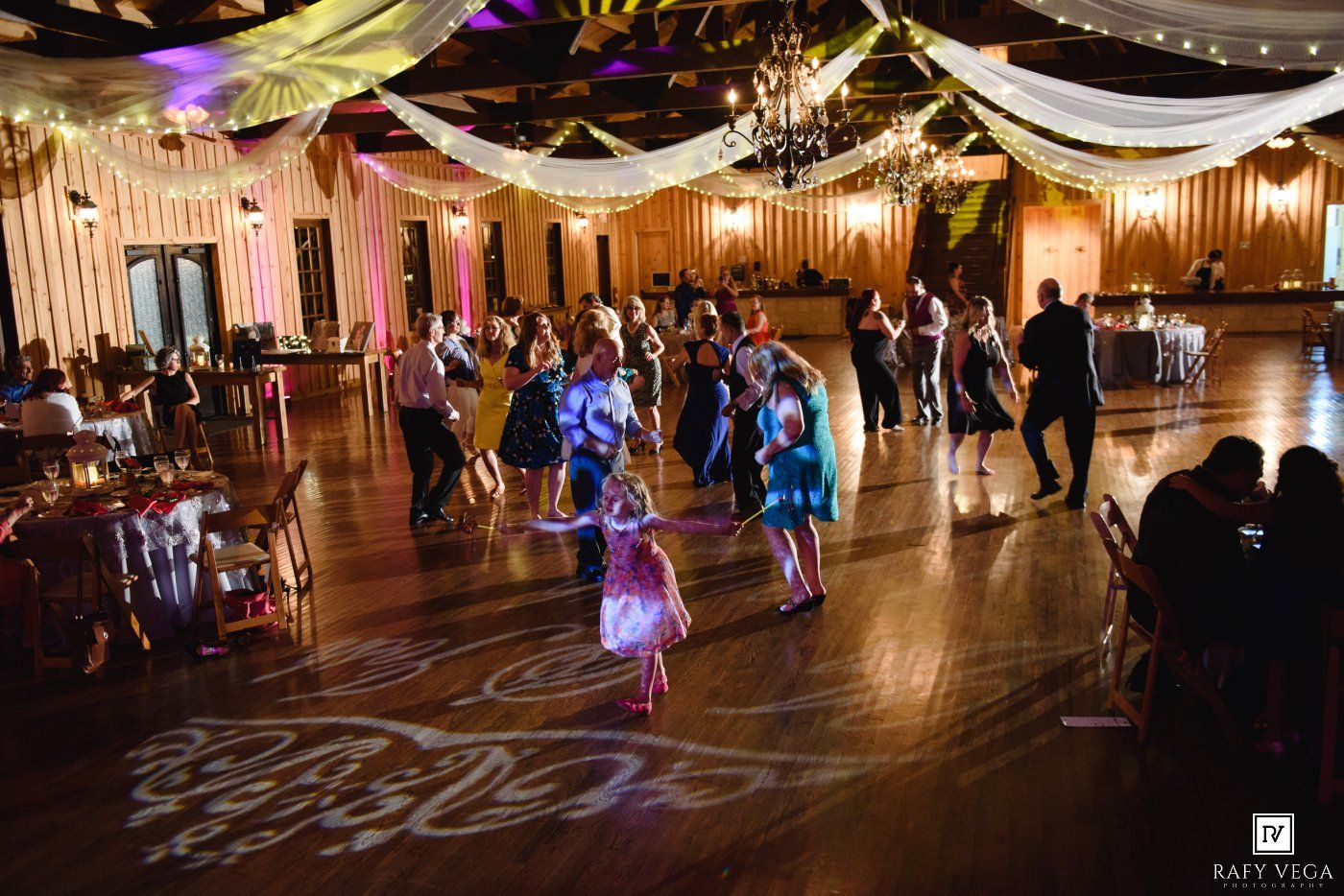 Guests dancing on a decorated ballroom floor at a wedding reception with colorful lights and elegant drapery.