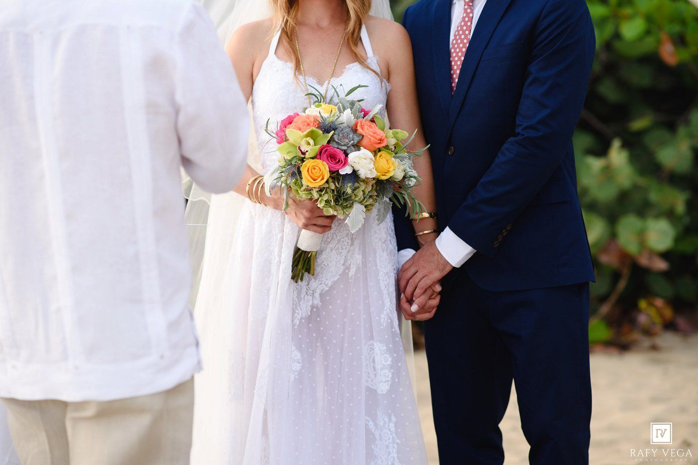 Bride in white dress holding colorful bouquet stands with groom in navy suit during outdoor beach wedding ceremony.