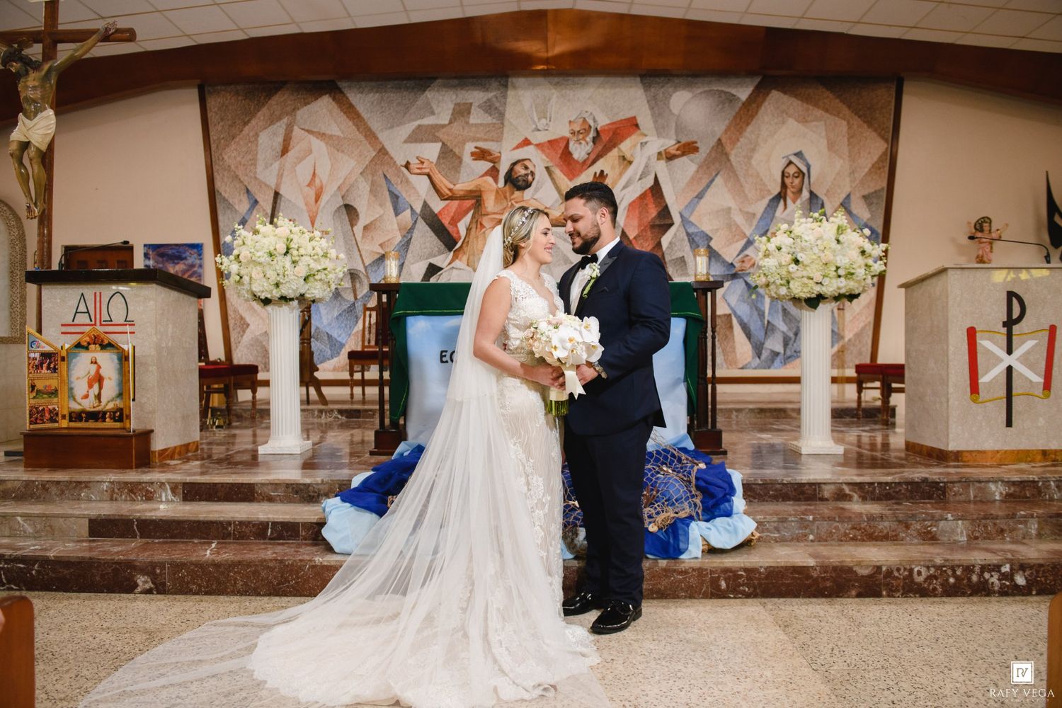 Bride and groom stand together at church altar adorned with white floral arrangements during wedding ceremony.