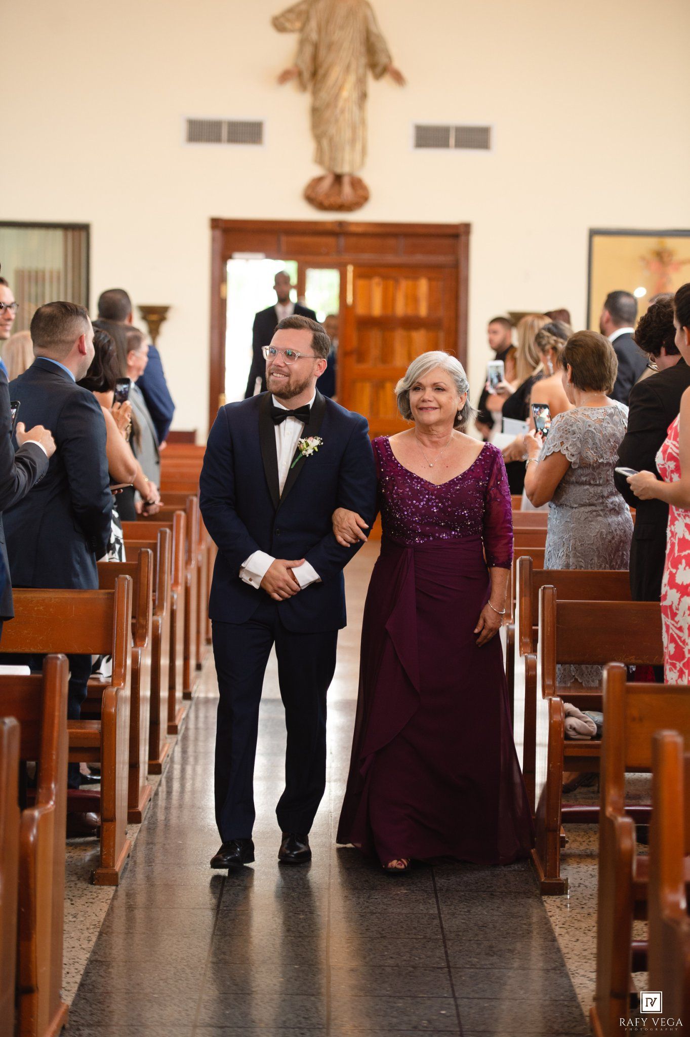 Groom in navy suit walks down church aisle with elderly woman in purple gown during wedding ceremony.