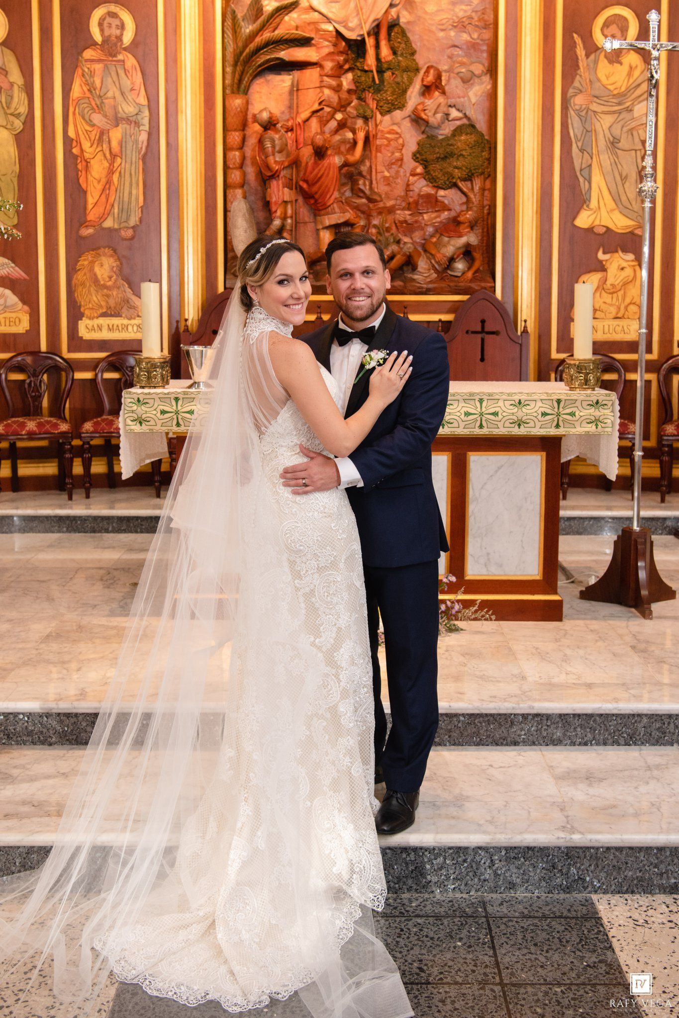 Bride in lace gown and veil poses with groom in navy suit at ornate church altar after wedding ceremony.