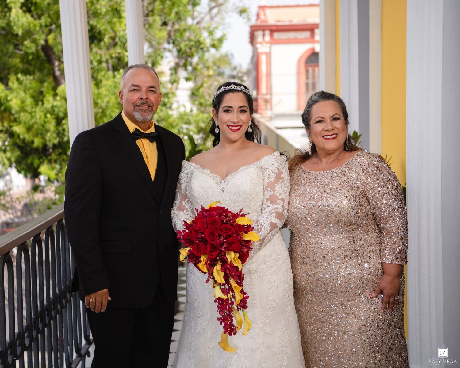 Bride in white gown holding red bouquet poses with parents on decorated balcony during wedding celebration.