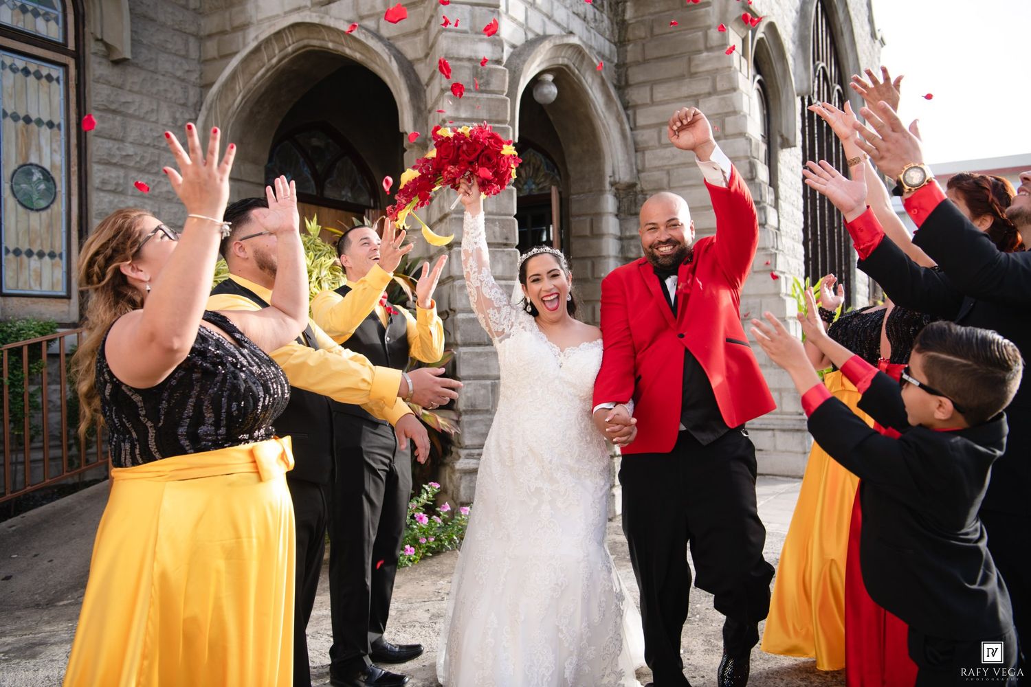 Bride and groom exit church as wedding party in yellow and red attire tosses rose petals in joyful celebration.