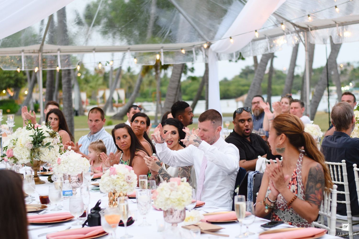 Guests celebrate at an elegant outdoor wedding reception under a white tent with floral centerpieces and string lights.