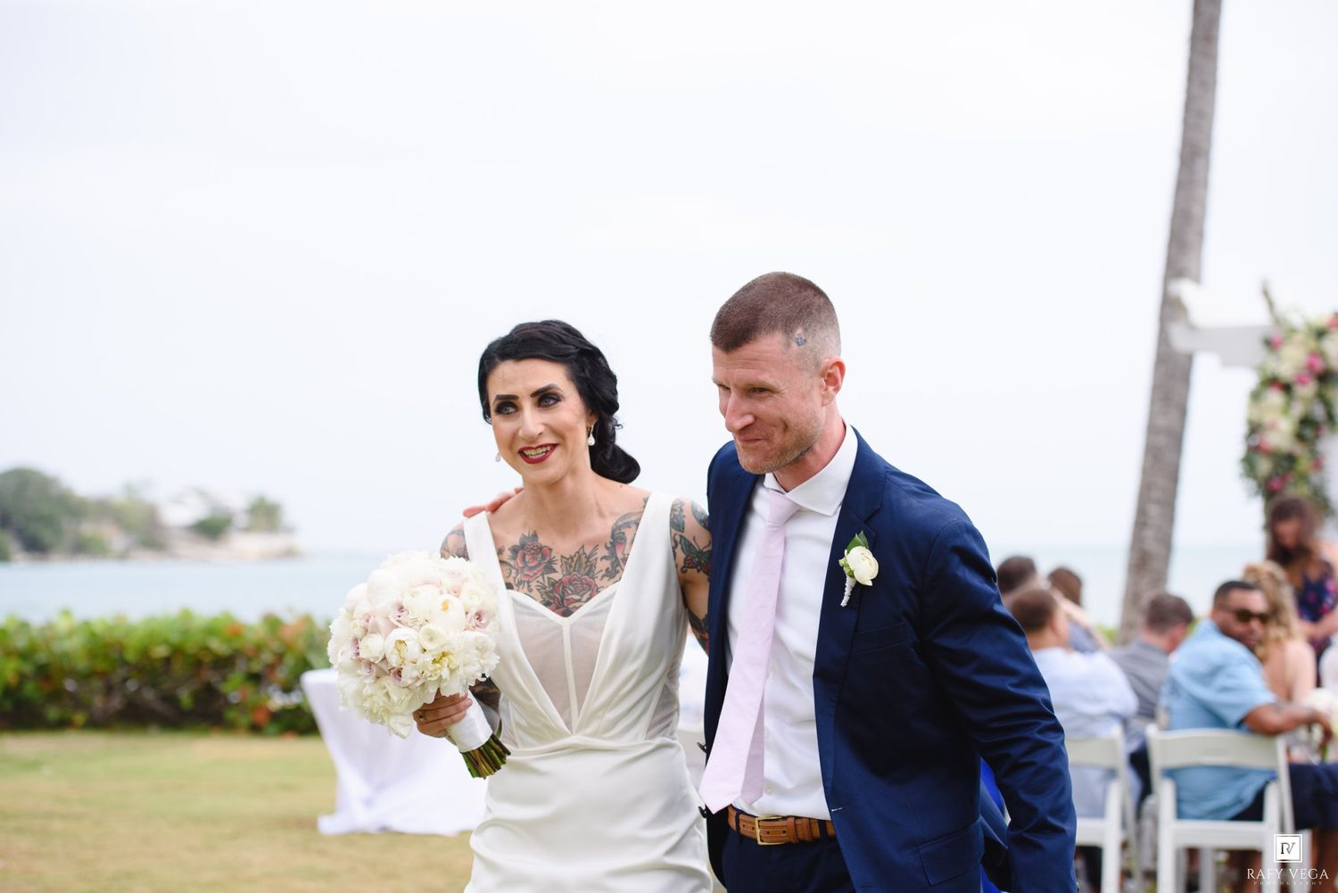 Smiling bride holding bouquet walks with groom after outdoor wedding ceremony near waterfront venue.