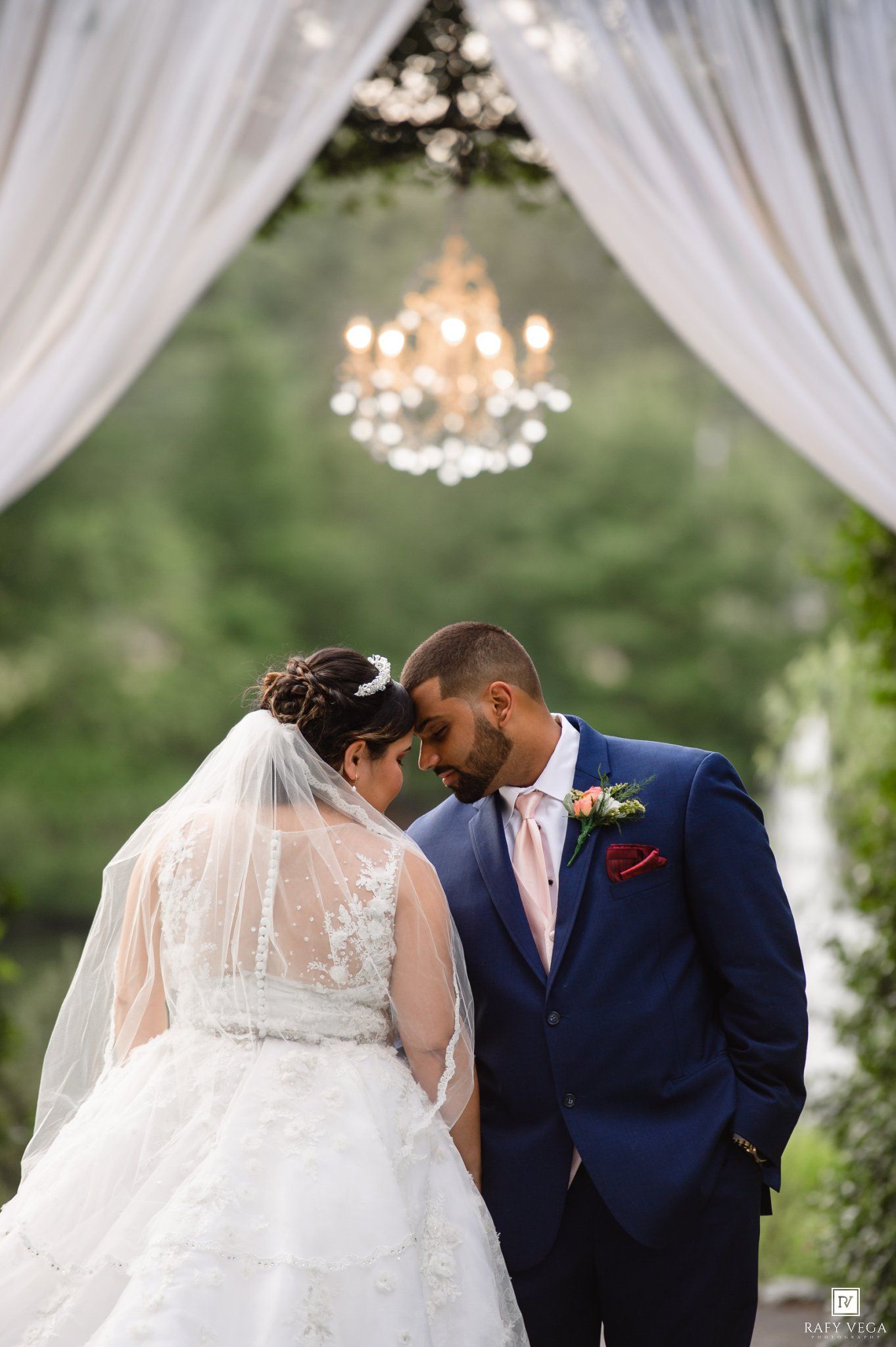 Bride and groom bow heads together under elegant chandelier and white draping at outdoor wedding ceremony.
