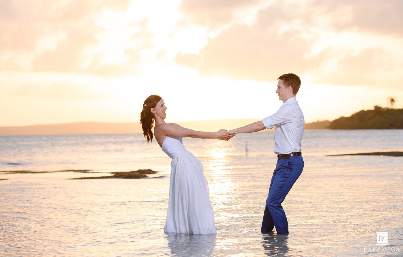 Couple dancing in shallow ocean water at sunset, woman in white dress holding hands with man in blue pants.
