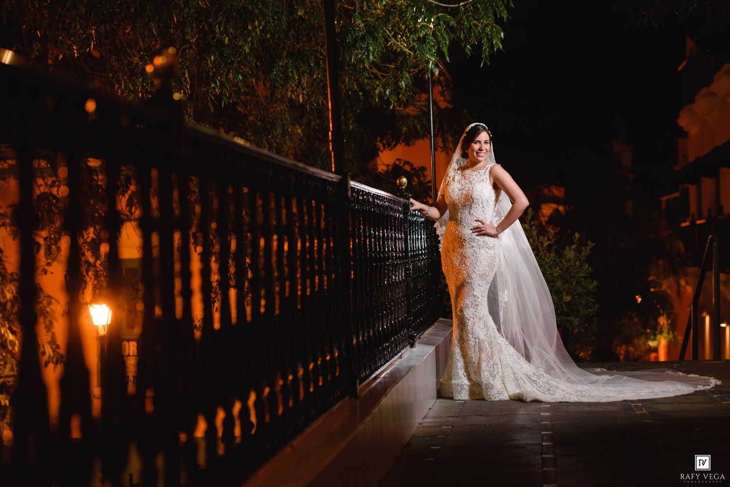 Bride in elegant lace wedding gown and veil poses on ornate bridge at night with warm golden lighting.