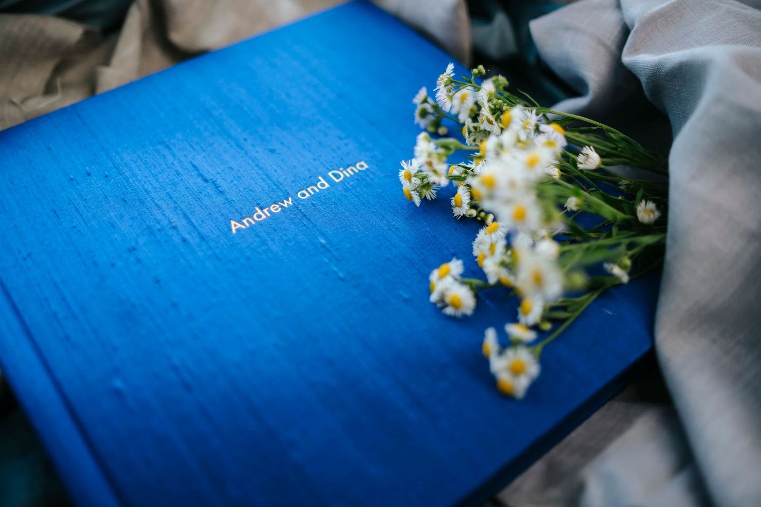 Blue wedding photo book titled Andrew and Dina adorned with white wildflowers on a soft gray fabric background.